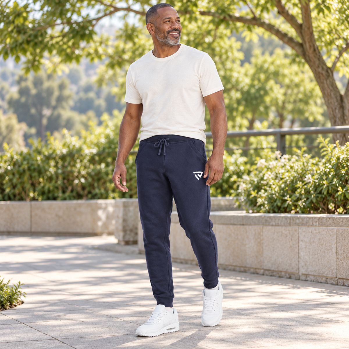 Men's True Fleece Joggers worn by an adult man in a clean modern park terrace with leafy trees in soft afternoon light; full-length view.